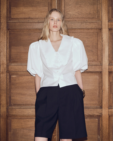 Woman wearing a white blouse and black shorts against a wooden paneled wall